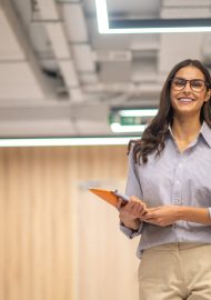 Good mood. Young long haired woman in glasses holding folder smiling at camera in illuminated room